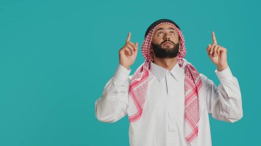 Orta Doğulu bir adam yukarıdan yukarıya bakıyor, stüdyoda yukarı doğru işaret ediyor. Geleneksel Arap kıyafetleri giyen genç İslamcı, başörtüsü ve cübbe giyiyor..