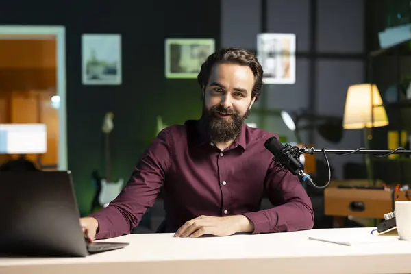 stock image Portrait of smiling man in studio looking at videos on laptop sent by viewers, filming with professional camera. Cheerful entertainer reacting to clips sent by audience during live broadcast