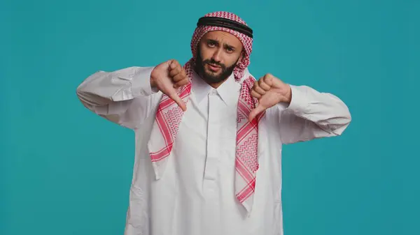 stock image In front of background, muslim adult gives thumbs up indicating fulfillment and happiness while dressed in traditional arabic clothes. Young man wearing religious islamic clothing, like sign.