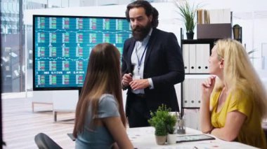 Finansal danışman müşterilerine pazar analizlerini geniş ekran kullanarak sunuyor ve veriye dayalı karar verme sürecini vurguluyor. Borsa göstergeleri hakkında istatistiksel çizelgeler gösteren broker, kamera A