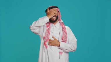 Smiling man laughing at jokes on camera, having fun dressed in muslim national clothes and kufiyah. Joyful arab adult enjoying laugh with friends, expressing happiness and positivity. clipart