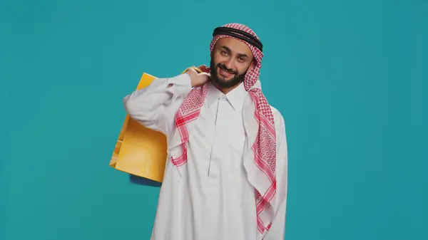 stock image Arab adult carrying shopping bags over blue background, posing with confidence after successful session of buying products. Young person in muslim clothes purchasing goods from mall on sale.