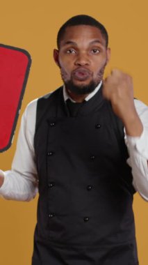 Dikey Video Garson siyaset için bir reklam yapıyor ve oy verme mesajıyla bir konuşma balonu gösteriyor, sarı arkaplana karşı duruyor. Restoran personelinde karton tabela var. Kamera B.