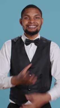 Dikey Video Güvenilir Neşeli Otel Kapıcısı stüdyoda kollarını kavuşturarak poz veriyor. İş yerinde smokin giyerken profesyonelliğini gösteriyor. Misafirperverlik sektörü çalışanı müşterileri selamlıyor