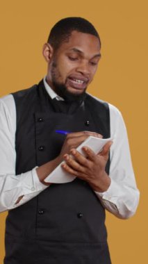 Vertical Video Professional Catering müşterilerine fatura için not defteri siparişleri yazıyor, sarı arka planda istenen yiyecekleri not alıyor. Önlüklü garson, müşteri memnuniyeti için defter servisi.