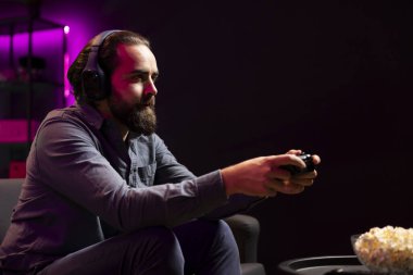 Man in neon lit apartment playing videogames on gaming console with teammates with bowl of popcorn in front. Player fighting foes in game, discussing with friends through headset and eating snacks clipart