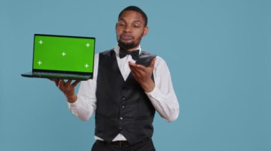 Kapıcı çocuk stüdyoda elinde yeşil ekranlı bir laptop tutarken beş yıldızlı bir otelde konukseverlik sektöründe çalışırken izole edilmiş krom anahtar görüntüsünü sunuyor. Profesyonel personel. Kamera B.