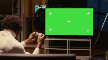 Man in dimly lit apartment playing video games on green screen widescreen smart TV, enjoying day off from work. Gamer battling enemies in online multiplayer shooter on gaming console clipart