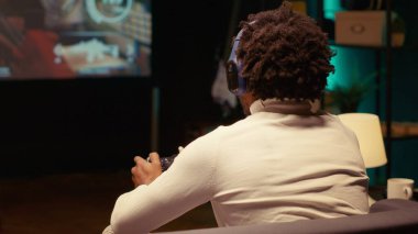 Close up shot of BIPOC man playing SF FPS videogame on widescreen smart TV, having fun in dark apartment room. Gamer competing in online multiplayer game, using headphones to chat with teammates clipart