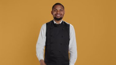 Garson gülümsüyor ve sarı arka planda baş parmak işareti gösteriyor, memnuniyetini ve onayını olumlu bir işaretle sunuyor. Neşeli çalışanlar iyi bir jest yapıyor. Kamera B.