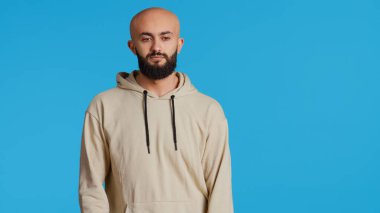 Ortadoğulu adam kamerada sıradan davranıyor ve havalı dans figürleri sergiliyor, bej kapüşonlu kapüşonlu rahat ve tasasız hissediyor. Genç yetişkin dansçı stüdyoda mavi arka planda eğleniyor. El kamerasıyla..