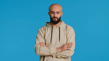 Kapüşonlu genç bir yetişkin stüdyoda bir şey bekliyor, kolları mavi arka planda çapraz duruyor. Ortadoğulu adam kamerada sabırsız, dakik ve gergin biri. Kamera 1.
