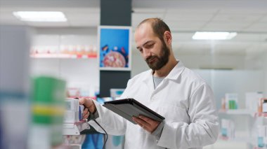 Eczane asistanı sağlık merkezindeki ilaç ve ilaç paketlerini tarıyor, raflardaki ilaç kaynaklarını izliyor. Çalışan tıbbi ürünleri izlemek için tablet ve tarayıcı kullanır.