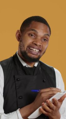 Vertical Video Professional Catering müşterilerine fatura için not defteri siparişleri yazıyor, sarı arka planda istenen yiyecekleri not alıyor. Önlüklü garson, müşteri memnuniyeti için defter servisi.