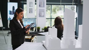 Proje planı ve kaba yazılım ile çalışan mimar, birlikte çalışma alanında 3D inşa modeliyle ölçümler alıyor. Mimarlık bürosu çalışanı mühendislik taslağına bakıyor..