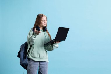 Sosyal medya bağlantılarını kontrol etmek için dizüstü bilgisayarı kullanan neşeli bir kadın ayakta duruyor. Tasasız genç, internet sitelerine not defteri cihazıyla bakıyor, mavi stüdyo arka planında izole edilmiş.