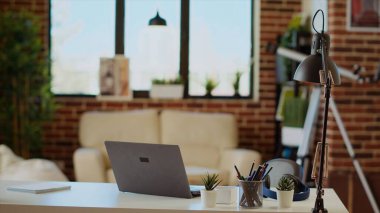 Büyük pencerelerden gelen güneş ışığıyla aydınlatılan rahat, boş bir daire, arka planda açık bırakılmış televizyon ve masa başında dizüstü bilgisayar. Tuğla duvarları ve eklektik iç dekorasyonu olan parlak modern oturma odası.