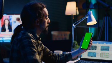 Elinde yeşil ekran akıllı telefonu olan bir telgraf çalışanı oturma odasındaki dizüstü bilgisayardaki istatistiksel çizelgeleri analiz ediyor. Serbest çalışan adam cep telefonundaki iş gelirlerini kontrol ediyor.