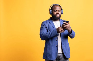 BIPOC 'lu adam en iyi arkadaşına akıllı telefon kullanarak mesaj atarken kulaklık takıyor. Afrikalı Amerikalı müzik dinliyor ve cep telefonundan arkadaşıyla sohbet ediyor, stüdyo arka planında izole edilmiş.