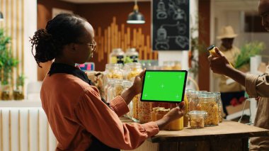 Afro-Amerikalı kadın süpermarkette doğal organik ürünlerin yanında duran tablet üzerinde yeşil ekran kullanıyor. Kadın satıcı elinde cihazla izole edilmiş bir fotokopi düzeneği gösteriyor. Kamera 1.