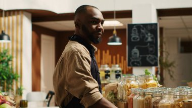 Önlüklü bir tüccar, eko dükkanındaki raflara büyük parçalar yerleştiriyor, sıfır atık konseptini ve sürdürülebilir yaşam tarzını destekliyor. Afrika kökenli Amerikalı satıcı ev yapımı kiler için reklam yapıyor. Kamera 1.