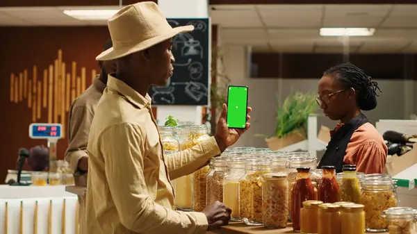 Stock image African american seller holding smartphone app with greenscreen, presenting isolated mockup template in local supermarket. Vegan merchant uses chromakey layout at organic grocery store. Camera 1.