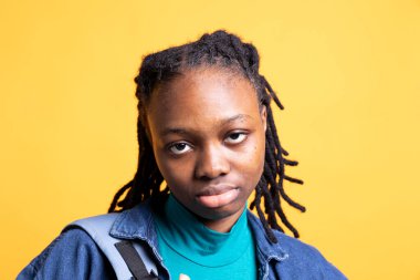 Poz veren kadın portresi, sarı stüdyo arka planında izole edilmiş, yakın çekim. Sıradan giyinmiş Afrikalı Amerikalı bir kız kameraya bakıyor, kendinden emin.