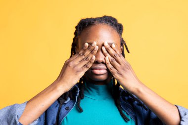 Bıkıp usanmış BIPOC 'lu genç kız yüzünü avuç içi ile kapatıyor. Okulda geçirdiği uzun bir günden sonra yorgun düşüyor. Kırılmaya ihtiyacı olan bir gencin yakın plan çekimi. Stüdyo arkaplanından izole edilmiş, elleriyle gözleri tıkayan.