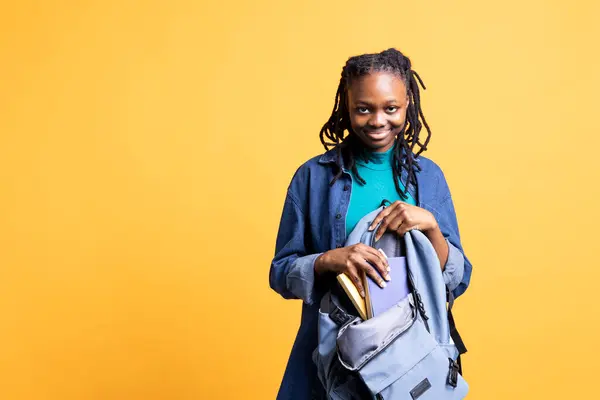 stock image Portrait of smiling teenager opening backpack to take encyclopedia books out for school project, studio background. Happy young girl removing textbooks from schoolbag, using them for academic purposes