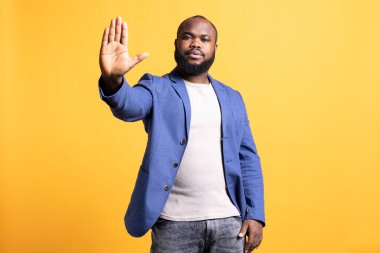 İddialı Afro-Amerikan adam dur işareti yapıyor. Sinirli BIPOC çalışanı dur işareti yapıyor, kavramı bitirmek istiyor, sarı stüdyo arka planında izole edilmiş.