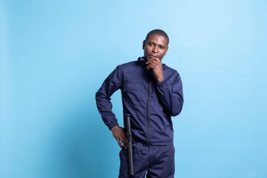 Üniformalı dalgın bir güvenlik görevlisi sorumluluğunu düşünüyor. Mülkü mavi arka plandan koruyor. Düşünceli, kararsız nöbetçi devriye görevinde çalışıyor..