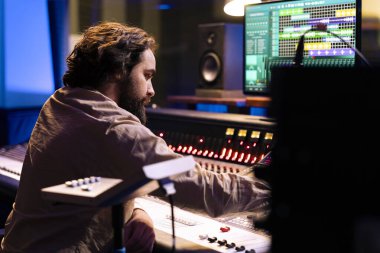 Ses teknisyeni stüdyoda yeni müzik kaydetme ve düzenleme üzerinde çalışıyor, kontrol odasında karıştırma ve çevirme düğmeleri kullanarak sesleri işliyor. Ses üreticisi düğmelere ve kaydırıcılara basar.