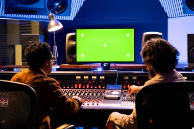 Profesyonel kayıt stüdyosunda müzik ve yeşil ekran montajı yapan müzisyenler kontrol odasındaki düğmeler ve faders ile ses karıştırma konsolu kullanıyorlar. Şarkılar besteleyen sanatçı ve yapımcı.