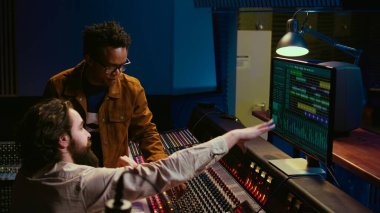Kayıt parçalarını ses yazılımıyla düzenlemede işbirliği yapan sanatçı takımı, ses seviyelerini ayarlamak için düğmeleri çeviriyor. Yapımcı ve müzisyen stüdyodaki kontrol masasında müzik yapıyor. Kamera B.