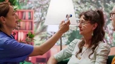 Tıbbi asistan, evdeki konsültasyon sırasında sıcaklık ve ateş ölçümlerini kontrol etmek için yaşlılar üzerinde termometre kullanıyor. Hemşire destek ve sağlık yardımı için kıdemli hastaları ziyaret ediyor. Kamera A.