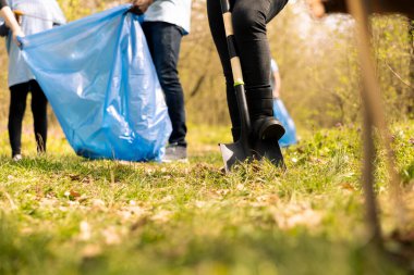 Eylemciler orman bölgesini çöp ve plastikten temizliyor, maşa ve poşetlerle çöp topluyorlar. İnsanlar doğal çevreyi korumak için gönüllü iş yapıyorlar, yasadışı çöplük..