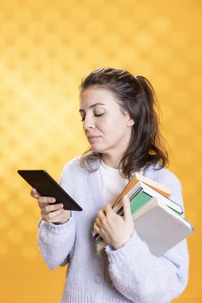 stock image Woman holding ebook reader and stack of books, isolated over studio background. Person with pile of textbooks in arms reading academic papers for research project on tablet