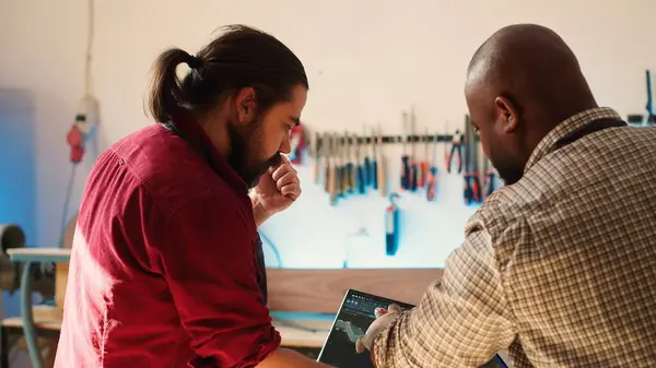 Stock image African american artist and colleague comparing wood art schematic with CAD software 3D render on laptop. Carpenters at work crosscheck drawing on clipboard with notebook blueprint, camera A