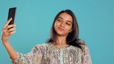Neşeli Hintli kadın akıllı telefon kullanarak selfie çekiyor ve sosyal medyada yayınlıyor. Mutlu insan fotoğraf çekiyor. Fotoğraf makinesi kullanıyor. Stüdyo arka planında izole edilmiş.