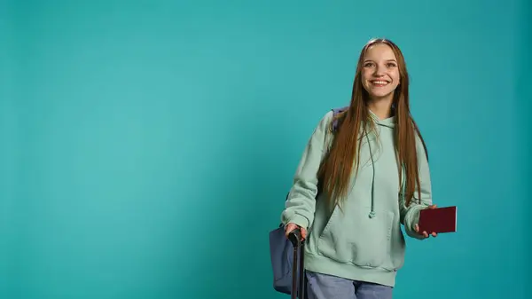 stock image Jolly teenage woman holding passport, going on adventure trip, looking around. Cheerful wanderlust tourist backpacking, sightseeing, visiting attractions, isolated over studio backdrop, camera B