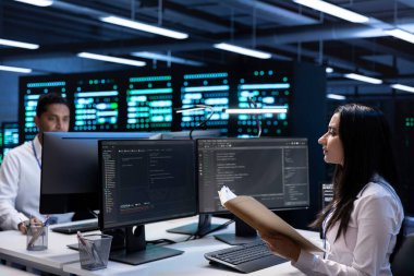 Veri merkezindeki mühendislerden oluşan bir ekip sistem arızasını önlemek için acil durum prosedürleri belgelerini kontrol ediyor. Sunucu çiftliği çalışma alanındaki meslektaşlar endüstri düzenlemelerine uyulmasını sağlıyorlar