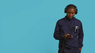 Security guard equipped with bodycam listening music, taking break from job shift. Portrait of bodyguard walking around perimeter with headphones on while at work, studio background, camera B clipart