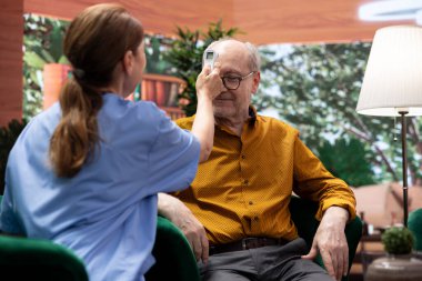 Tıbbi asistan, evdeki konsültasyon sırasında sıcaklık ve ateş ölçümlerini kontrol etmek için yaşlılar üzerinde termometre kullanıyor. Hemşire destek ve sağlık yardımı için kıdemli hastaları ziyaret ediyor.