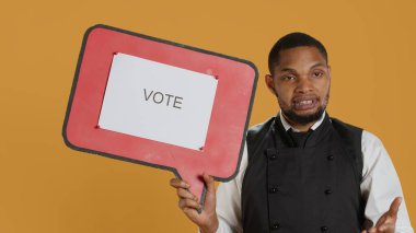 Professional waiter showing a message to vote on speech bubble, creating an advertisement for politics on cardboard icon. Restaurant employee working in customer oriented industry. Camera A. clipart