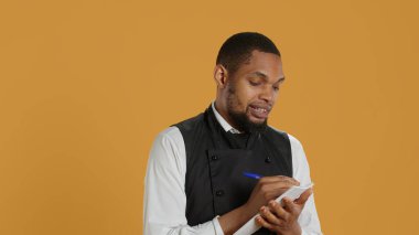 Profesyonel yemek şirketi müşterilerine fatura için not defteri hazırlıyor. Sarı arka planda istenen yiyecekleri not alıyor. Önlüklü garson, müşteri memnuniyeti için defter servisi. Kamera A.