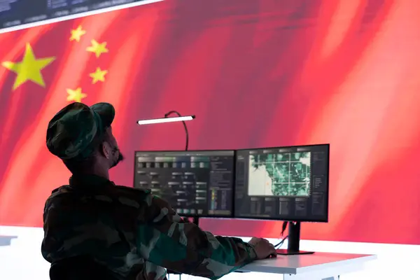 stock image Chinese military officer in camouflage uniform monitors tactical information on computer. Soldier gathering intelligence via radar surveillance and gps, national flag of China in control center.