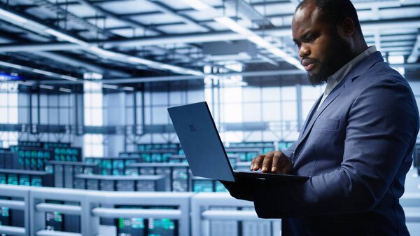 Software developer in server hub checking recovery plan on laptop, monitoring hardware energy consumption. African american worker uses notebook in data center, ensuring rigs are functioning, camera B