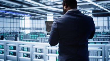 Veri merkezindeki Afrikalı Amerikalı yönetici yazılım hatalarını düzeltmek için tablet üzerindeki araçları kontrol ederken hata yaptı. Sunucu merkezi çalışma alanı bilişim uzmanı ana bilgisayarların performansını etkileyen sorunları arıyor, kamera B