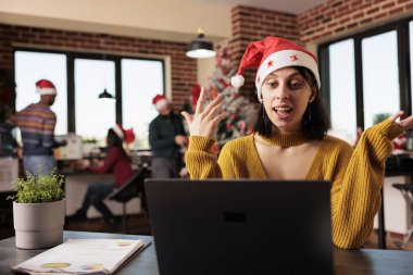 Kafkas kadın proje yöneticisi Noel süslemeli ofiste dizüstü bilgisayarla toplantıya katılıyor. Noel Baba şapkalı şirket çalışanı kış tatilinde video çağrısına başlama konusunu tartışıyor.