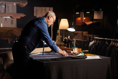 Yaşlı titiz takım elbiseli adam profesyonel terzilik stüdyosunda kaliteli giysiler üzerine ölçümler yapıyor. Atölyede kıdemli moda tasarımcısı yaklaşan giysi koleksiyonu üzerinde çalışıyor.